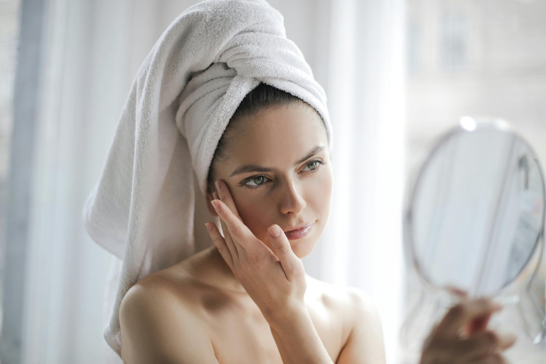 A woman in a white robe and towel performs her K-Beauty skincare ritual, applying a moisturizer to her cheek while looking in the mirror to observe her skin type.